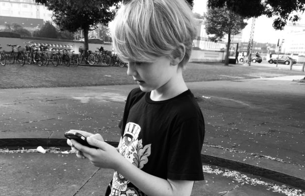 Dreng med lyst hår og en mobiltelefon i hænderne . står i byen han kigger på telefonen